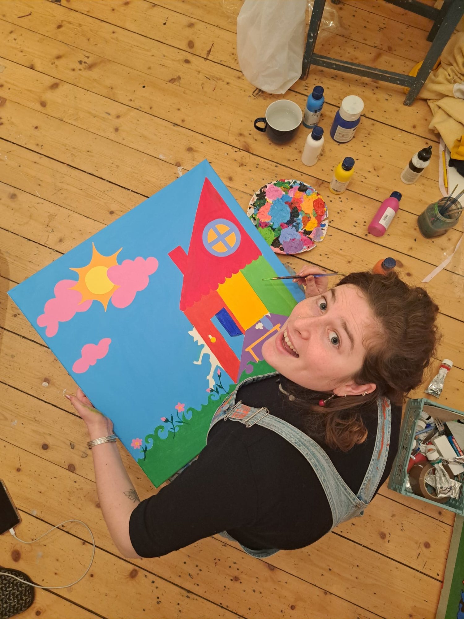 Maria seen from above, painting a canvas of a red-roofed house on the studio floor